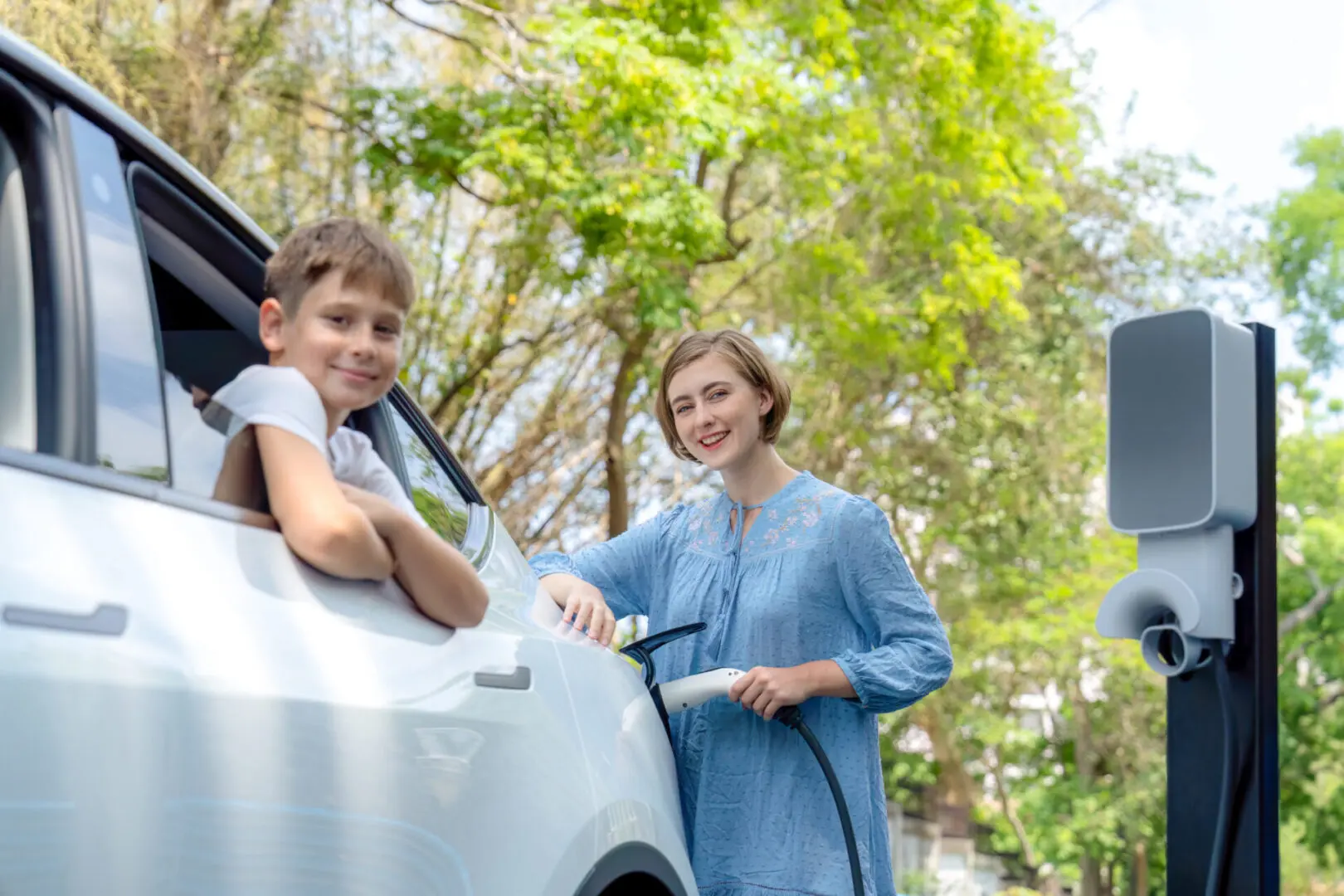 A smiling woman and boy standing by a car under green trees.
