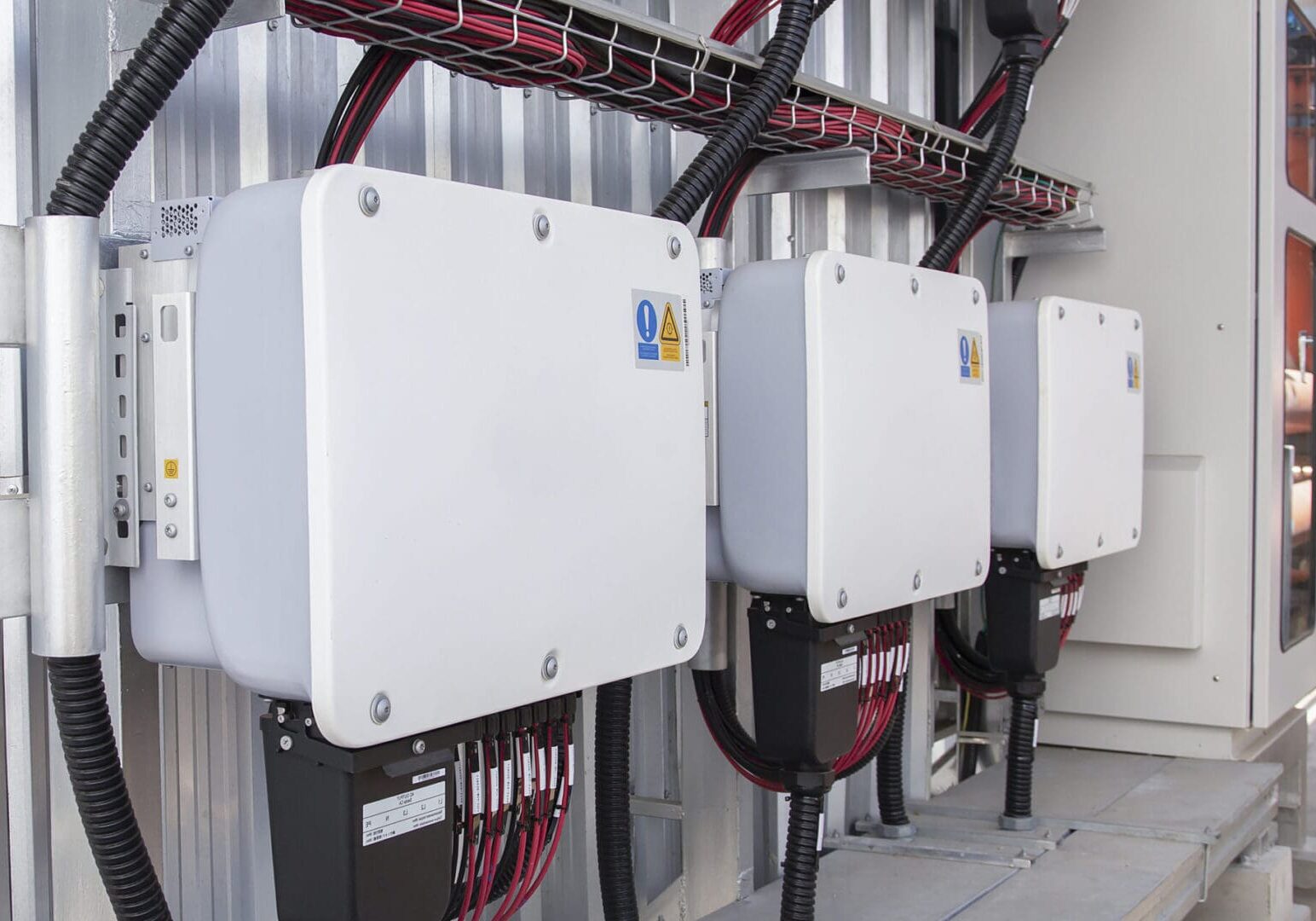 Three white electrical inverter units mounted on a wall with connected cables.
